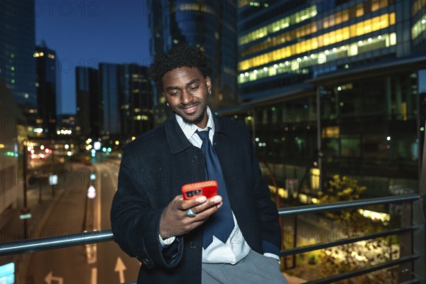 Young black businessman on an elevated walkway at night, smiling as he uses his smartphone with blurred city lights and modern buildings behind, conveying urban tech and connection
