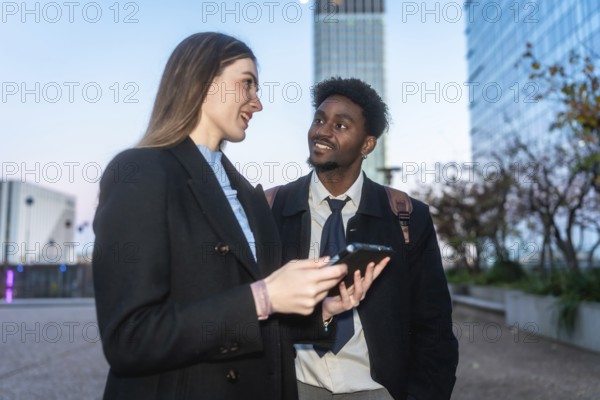 Two diverse business colleagues engaging in a discussion and sharing information on a digital tablet, standing outdoors in a modern urban environment with office buildings