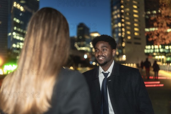 Young african american man engaging in a business conversation with a female colleague outdoors, with blurred city lights and buildings providing a dynamic urban backdrop during the evening