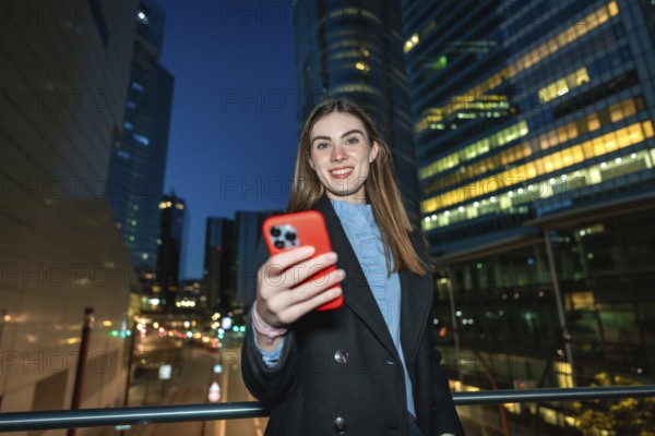 Young woman smiling and holding a smartphone, capturing a selfie or video while standing on a bridge in a modern city at night with illuminated skyscrapers in the background