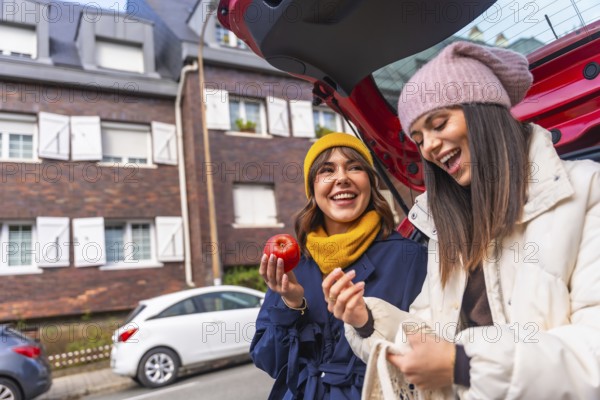 Two cheerful young multiracial women enjoying a moment of laughter while unloading fresh produce, including a red apple, from a car trunk after a successful grocery shopping trip