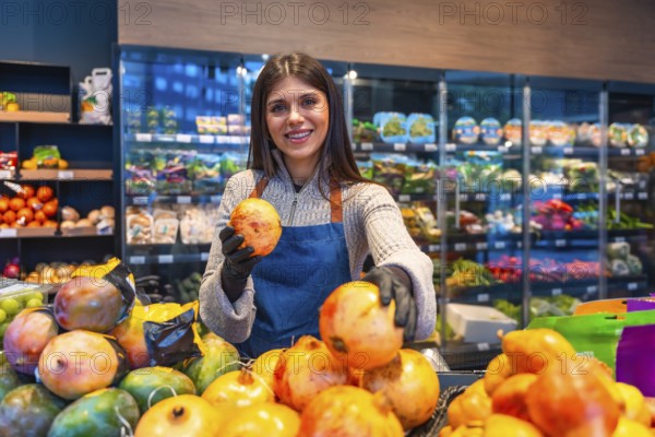 Smiling young woman greengrocer wearing an apron and gloves at a supermarket produce stand, proudly presenting fresh pomegranates to customers, ensuring quality and excellent service