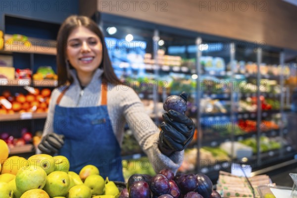 Smiling saleswoman in an apron and gloves working at a supermarket's produce section, holding a fresh plum while surrounded by various fruits and vegetables