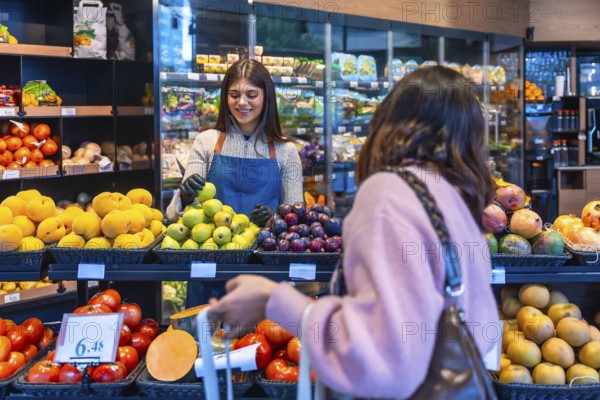 Friendly supermarket employee wearing an apron and gloves assisting a female customer in the produce section by offering a fresh green apple, highlighting good service and healthy food choices