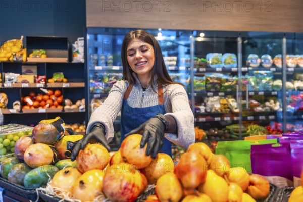 Smiling young supermarket employee in apron and gloves arranging fresh pomegranates, mangos and grapes on produce display, highlighting quality, care and healthy retail service