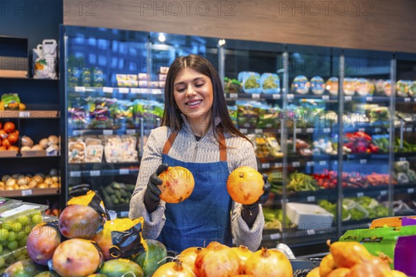 Female grocery store employee wearing an apron and gloves, smiling while holding two pomegranates, arranging healthy fruit for fresh consumption on a display shelf