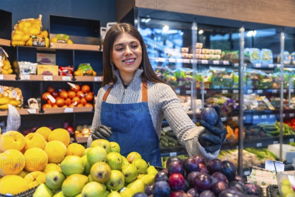 Greengrocer woman smiling as she arranges vibrant fresh plums and assorted produce at a market counter, offering quality, variety and friendly customer service