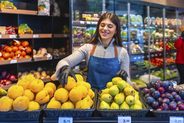 Woman grocer wearing gloves and an apron, smiling while arranging fresh produce like apricots, pears, and plums on display in a modern supermarket or grocery store