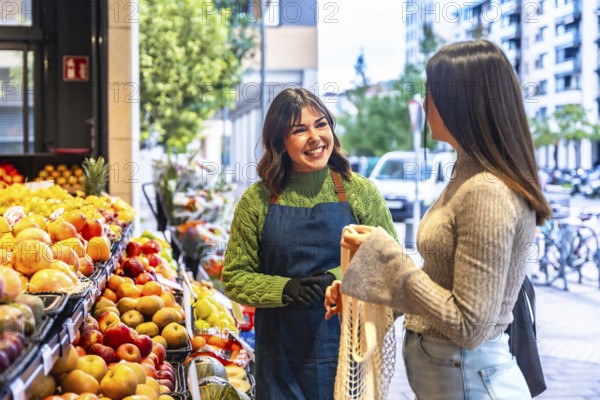Smiling greengrocer wearing an apron and gloves receiving a female customer holding a reusable net bag to buy fresh and healthy organic fruits and vegetables outside a small market