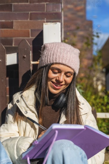 Young woman in a beanie and headphones writing in a notebook outdoors on a sunny day, smiling and focused as she journals, studies ideas, and enjoys a peaceful moment