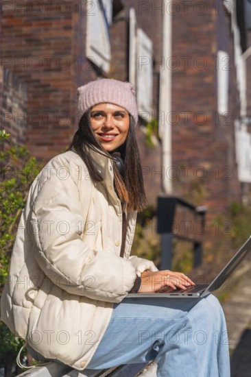 Woman in a cozy winter hat and jacket sitting outdoors, smiling while typing on a laptop, enjoying remote work and connectivity in an urban environment