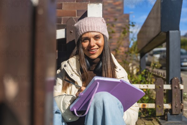 Young woman in beanie and headphones smiles while sketching in a notebook on a sunny urban street, relaxed and focused, enjoying creative outdoor downtime