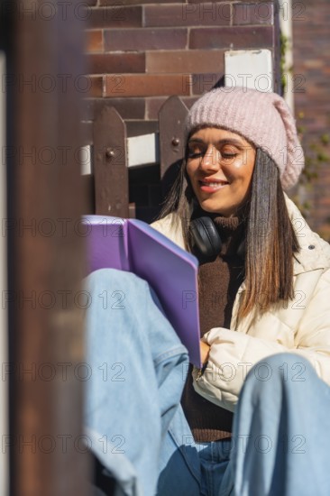 Woman smiling with closed eyes, wearing a beanie and headphones, sitting peacefully outdoors in the warm sunlight while reading a book, finding joy and relaxation