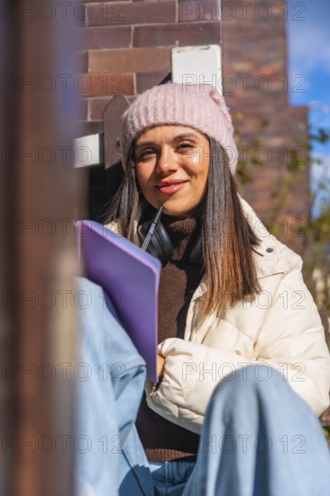 Woman student smiling and looking at the camera while writing in a journal outdoors, wearing a pink beanie, a puffer jacket, and headphones, studying and learning on a sunny day