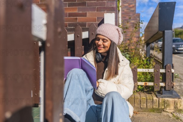 Young woman sitting on the sidewalk enjoying a good book, dressed in a warm winter coat and beanie, with headphones around her neck, smiling while immersed in reading under the sunlight