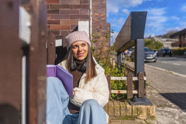 Young woman sitting outdoors on a sunny day, wearing a beanie and headphones, holding a book, looking away thoughtfully while enjoying a moment of relaxation and study