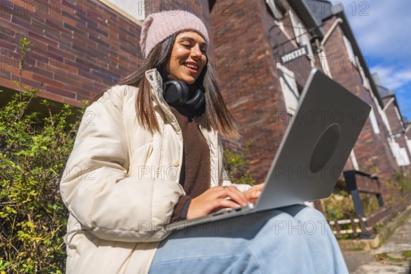 Young woman smiling and typing on a laptop, dressed in winter clothes, sitting outside a brick building on a sunny day, representing remote work and digital nomad lifestyle