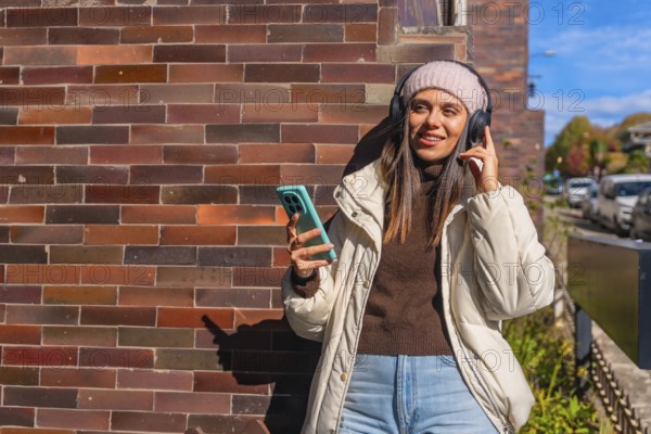 Young woman leaning against a brick wall, smiling with wireless headphones and smartphone, enjoying music and sunny urban street life on a casual autumn day