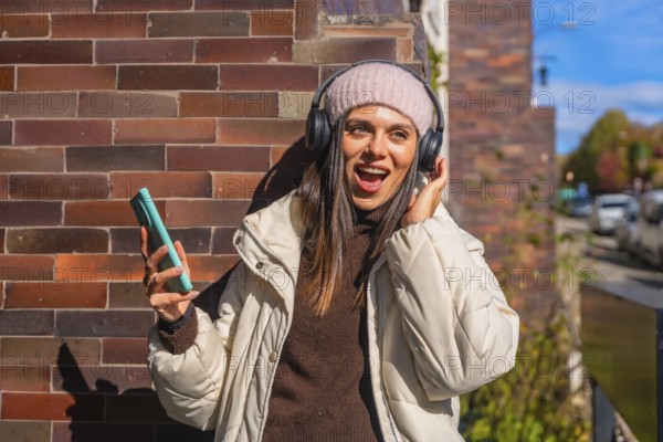 Young woman singing along to music on wireless headphones, holding her phone against a brick wall in a vibrant autumn city scene, smiling and enjoying the moment