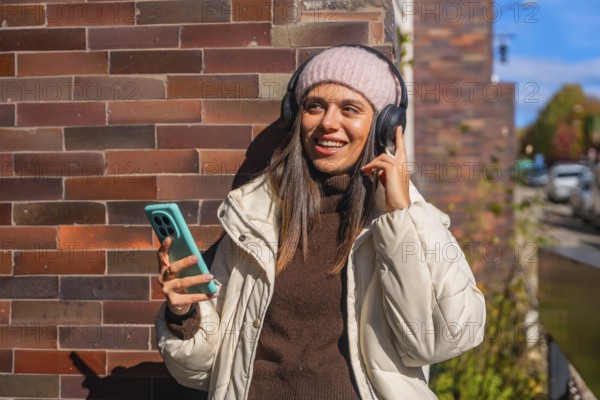 Young woman smiling while enjoying music through her headphones, holding a smartphone outdoors against a brick wall on a sunny autumn day, embodying relaxation and urban lifestyle