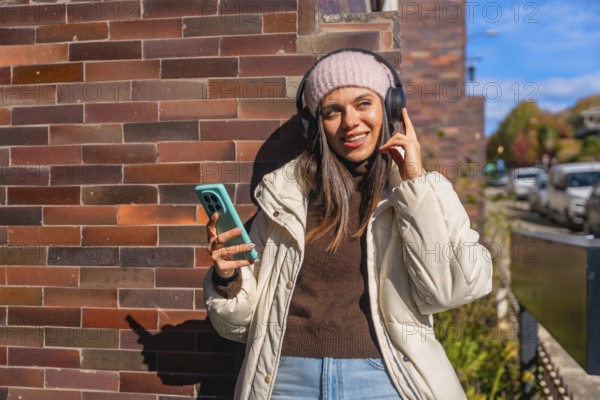 Smiling young woman dressed in warm winter clothes standing by a brick wall, peacefully listening to an audio using her wireless headphones and mobile phone under bright sunlight