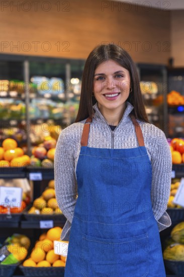 Young woman wearing a denim apron and a sweater, smiling confidently while standing in a supermarket aisle with fresh produce on display, representing service and retail