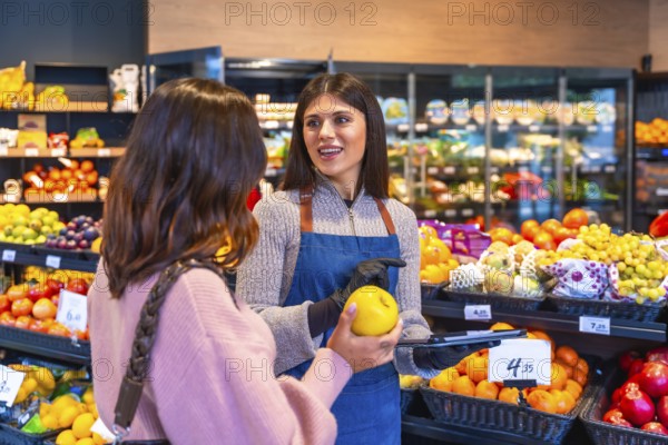 Supermarket employee wearing an apron and gloves helping a woman consumer select a fresh yellow apple from the fruit section, talking about product quality and service