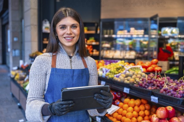 Greengrocer woman wearing an apron and gloves, standing in a small business produce shop, holding a digital tablet, managing inventory with fresh fruits and vegetables around her