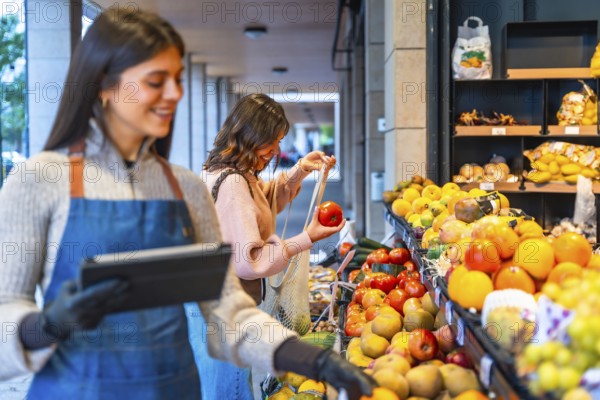 Greengrocer smiling and holding a digital tablet while a happy woman consumer chooses fresh tomatoes, fruits, and vegetables from an outdoor market stall, promoting healthy eating and local business