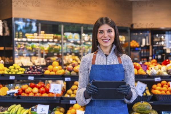Smiling woman wearing an apron and gloves handling a digital tablet in a grocery store, representing retail business management, customer service, and fresh organic produce