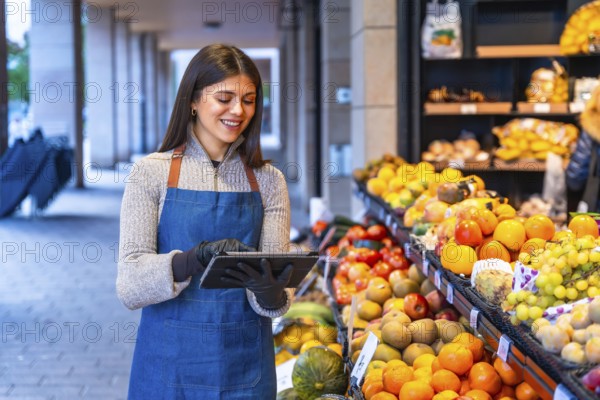 Smiling woman grocer in apron and gloves uses a tablet to manage inventory and orders at a fresh produce stand, modern small business efficiency and customer service