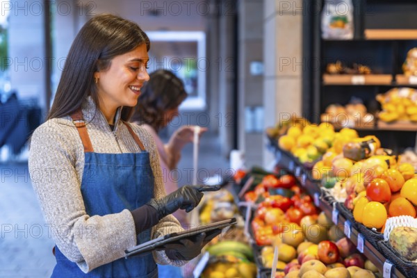 Female shop assistant wearing an apron and gloves managing fresh fruit and vegetables inventory with a digital tablet in a local grocery store, smiling while working