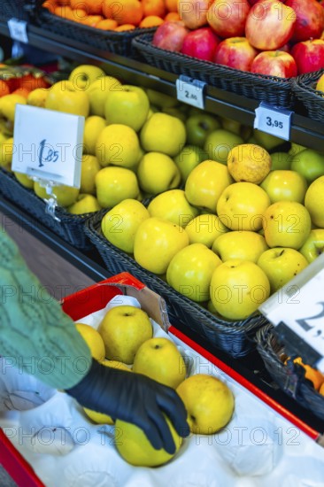 Person wearing a black glove carefully choosing ripe yellow apples from a display case at the fresh produce section in a supermarket, promoting healthy eating and conscious shopping choices