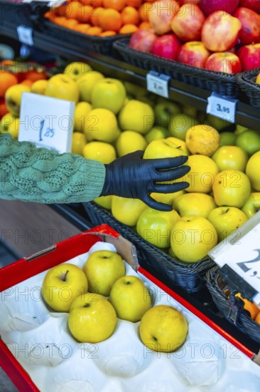 Person's hand, wearing a black protective glove, selecting fresh golden delicious apples from a display in a supermarket, alongside other colorful fruits