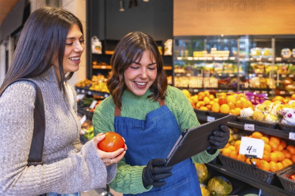 Store employee in an apron and gloves using a tablet while helping a smiling customer holding a fresh tomato in the vibrant produce section of a modern grocery store
