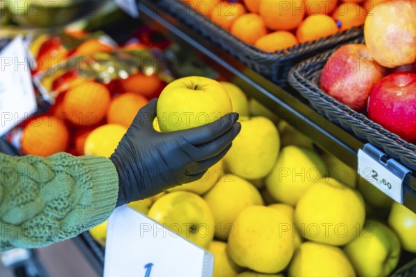 Hand in a black protective glove selects a fresh yellow apple from a supermarket produce display, highlighting hygiene, safety and responsible shopping in the store aisle