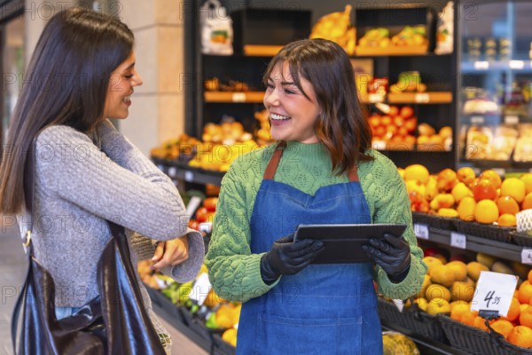 Friendly store employee wearing an apron and gloves holding a digital tablet, helping a smiling female customer with her shopping in the brightly lit produce aisle of a supermarket