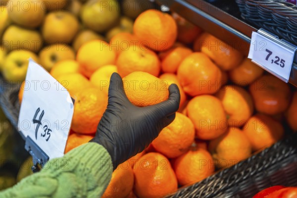 Person wearing a protective glove picking an orange from a large display of citrus fruits in a grocery store, demonstrating safe public shopping practices during a pandemic