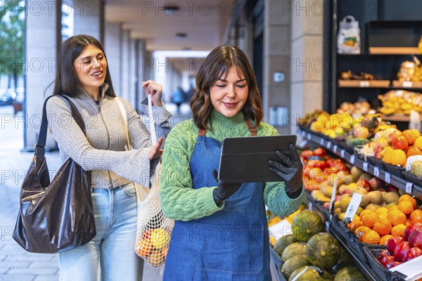Greengrocer in apron and gloves uses a tablet to manage inventory and assist a smiling customer selecting fresh fruits and vegetables at an outdoor market stall