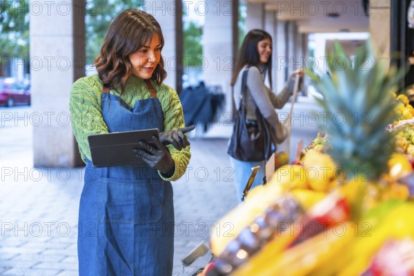Female small business owner wearing an apron and gloves managing inventory and sales on a digital tablet at her fresh produce stall while a customer shops in the background