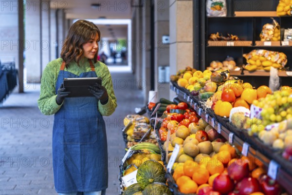 Woman grocer using a digital tablet to manage inventory and fresh produce at her outdoor fruit and vegetable stand, highlighting modern technology in small business retail