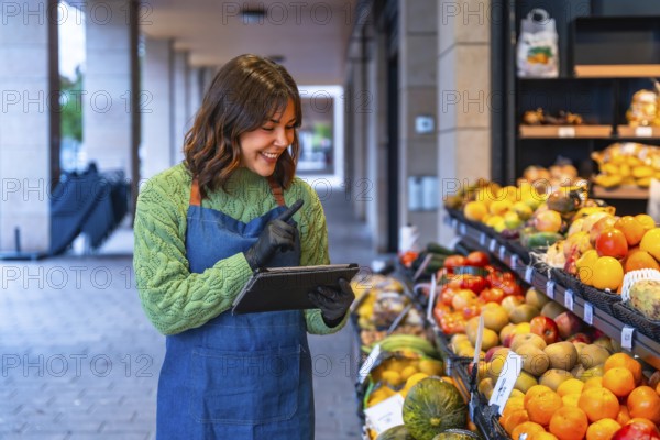 Woman in an apron and gloves managing fresh produce at her small business, using a digital tablet for inventory control while standing next to vibrant displays of fruit and vegetables