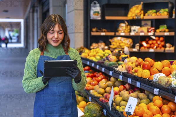Female small business owner wearing an apron and gloves managing stock and orders on a digital tablet while standing in front of a colorful fruit and vegetable display