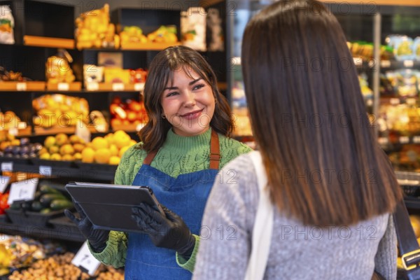 Friendly saleswoman holding a tablet and wearing gloves, assisting a customer with produce in a modern supermarket while offering excellent service and support