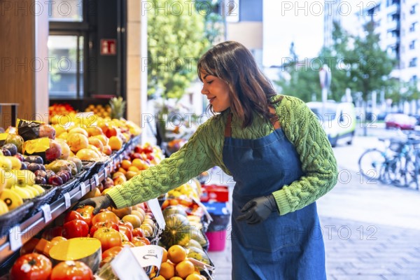 Smiling young greengrocer woman wearing an apron and gloves arranging fresh fruit and vegetables on market stalls outside her small business, ensuring quality and presentation