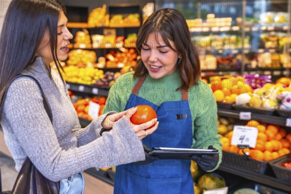 Grocery store employee in an apron holding a tablet and helping a female customer examine a fresh tomato in the produce aisle, fostering excellent customer service and shopping experience