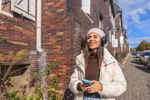 Smiling young woman walking through a sunny urban street in autumn, listening to music on headphones and enjoying a bright, carefree moment with her phone in hand