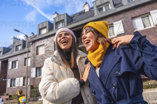 Two cheerful young women friends enjoying a sunny city day, laughing wholeheartedly while wearing warm winter clothing and walking arm in arm, symbolizing genuine happiness and strong friendship