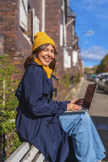 Young woman smiling at camera while sitting on a public bench in an autumn street, typing on her laptop, embodying the concept of flexible remote work and a digital nomad lifestyle