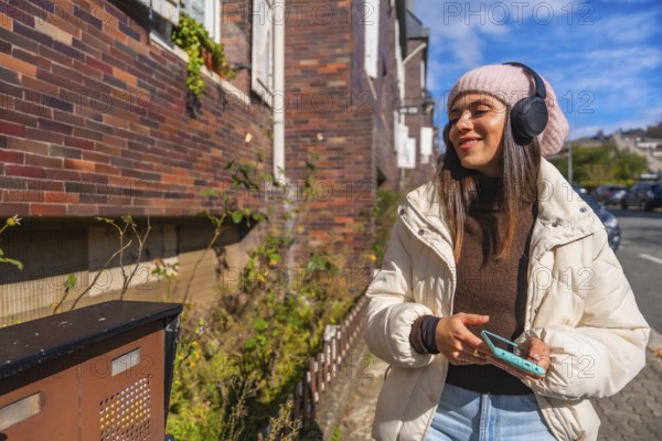 Young woman smiling and listening to music through headphones while walking outdoors on an urban street, dressed in warm clothes and holding a smartphone on a sunny autumn day